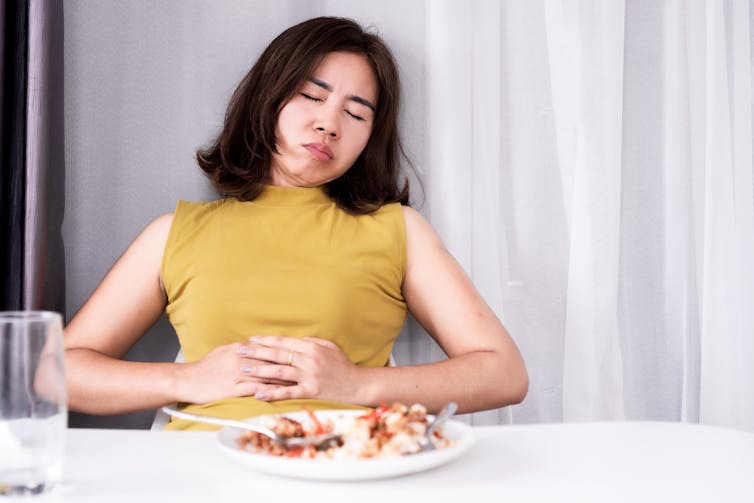 A woman leans back from her plate at the table holding her stomach because she’s eaten too much.